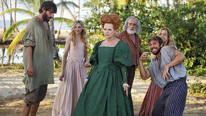 En una playa con palmeras, un grupo de personas vestidas con ropa de época interactúan. Una mujer con vestido verde y peinado rojizo destaca, mientras otros personajes observan y reaccionan a la escena.