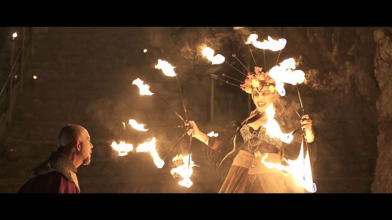 Una artista femenina, con un tocado de flores y un vestido detallado, manipula varas de fuego en un entorno oscuro, posiblemente un espacio cerrado. Un hombre observa la actuación.
