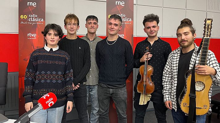 Seis personas posan en un estudio de radio, con micrófonos y un panel con logos al fondo. Los individuos visten ropa casual, incluyendo suéteres y camisas, y algunos sostienen instrumentos musicales.