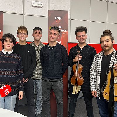 Seis personas posan en un estudio de radio, con micrófonos y un panel con logos al fondo. Los individuos visten ropa casual, incluyendo suéteres y camisas, y algunos sostienen instrumentos musicales.