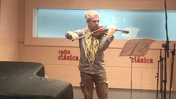 En un estudio de grabación, un violinista con camisa gris y bufanda amarilla interpreta una pieza musical. Un atril con partituras y un piano de cola negro complementan la escena, con el logo de la emisora en el fondo.