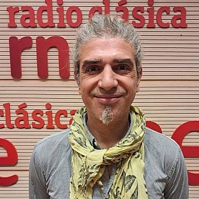 En un estudio de grabación, un violinista con camisa gris y bufanda amarilla interpreta una pieza musical. Un atril con partituras y un piano de cola negro complementan la escena, con el logo de la emisora en el fondo.