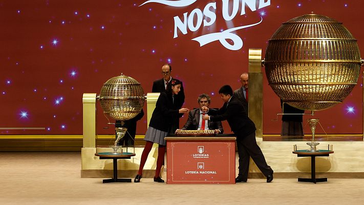 En el escenario del sorteo, dos bombos dorados se alzan sobre un fondo rojo oscuro con estrellas. Una mesa roja con el logo de \"Loterías\" y un hombre con traje y corbata roja son elementos centrales, junto a niños y adultos que participan en el evento.