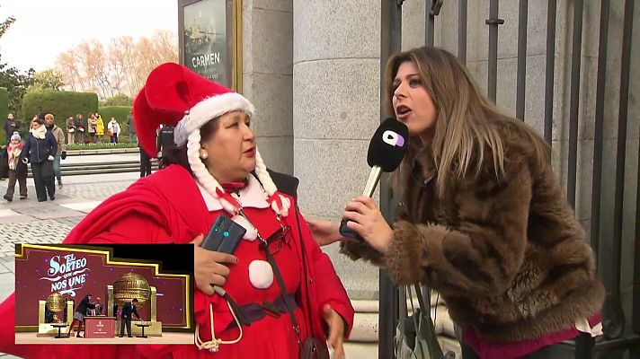 Una reportera con un micrófono entrevista a una mujer disfrazada de Mamá Noel. En el teléfono de la mujer disfrazada se muestra una imagen de un sorteo de lotería.