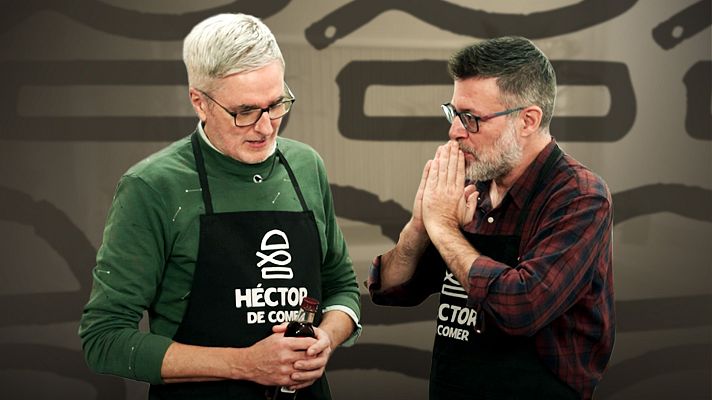 Dos hombres en un estudio de cocina, ambos con delantales negros con el texto \"HÉCTOR DE COMER\", posan para la cámara. Uno sostiene una botella y el otro se lleva las manos a la boca.
