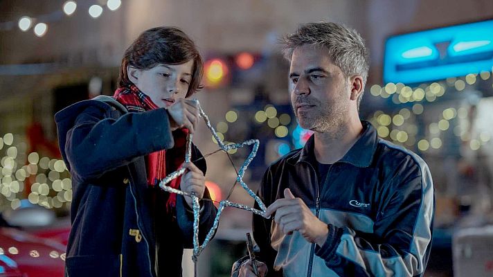 Un niño y un hombre decoran una estrella navideña. El niño, con bufanda a cuadros, trabaja en la estrella de alambre, mientras el hombre observa. Luces navideñas borrosas sugieren un ambiente festivo.