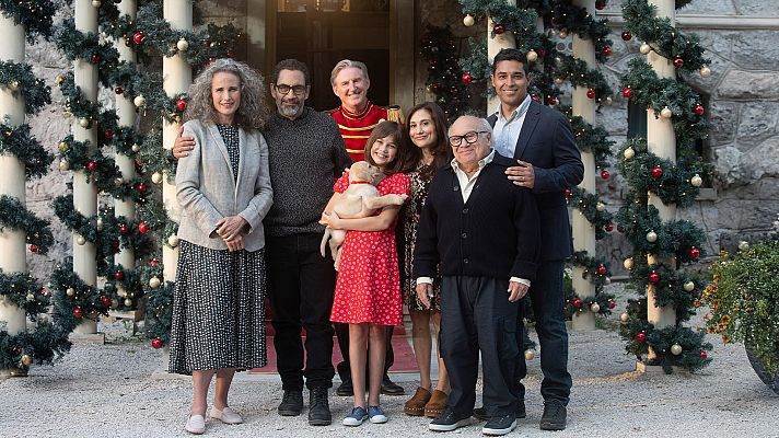 Un grupo de personas sonríe frente a un edificio decorado con adornos navideños, con guirnaldas y luces. Una niña sostiene un cachorro, y entre ellos se encuentra un hombre reconocido.