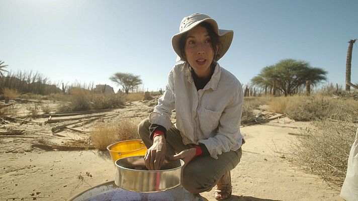 En un entorno desértico, una persona arrodillada utiliza un tamiz metálico, con un cubo amarillo cerca. La escena forma parte de una expedición documental, con vegetación escasa y árboles dispersos.