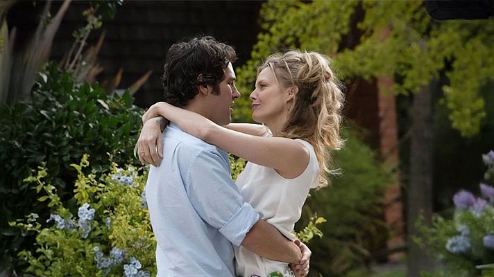 Una pareja se abraza en un entorno natural; el hombre con camisa azul y cabello oscuro, la mujer con vestido blanco y cabello rubio recogido, se miran con afecto, rodeados de vegetación.