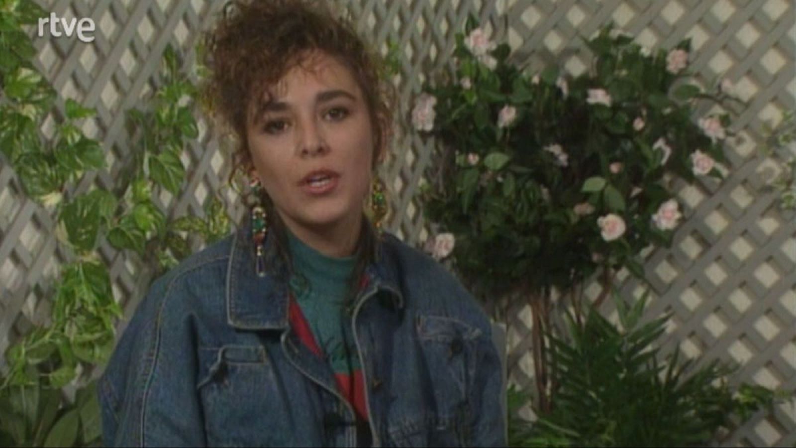 Isabel Gemio, presentadora de "Hablemos de sexo" en RTVE, con cabello rizado, pendientes llamativos, camiseta teal y chaqueta vaquera, posa ante un fondo de jardín con el logo de RTVE.
