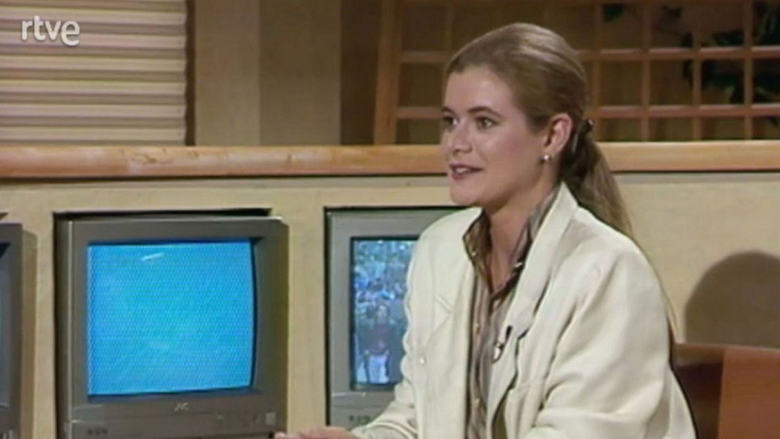Elena Ochoa en un estudio de RTVE, con monitores CRT antiguos y una estructura de celosía de madera al fondo. Viste chaqueta clara y camisa de rayas.