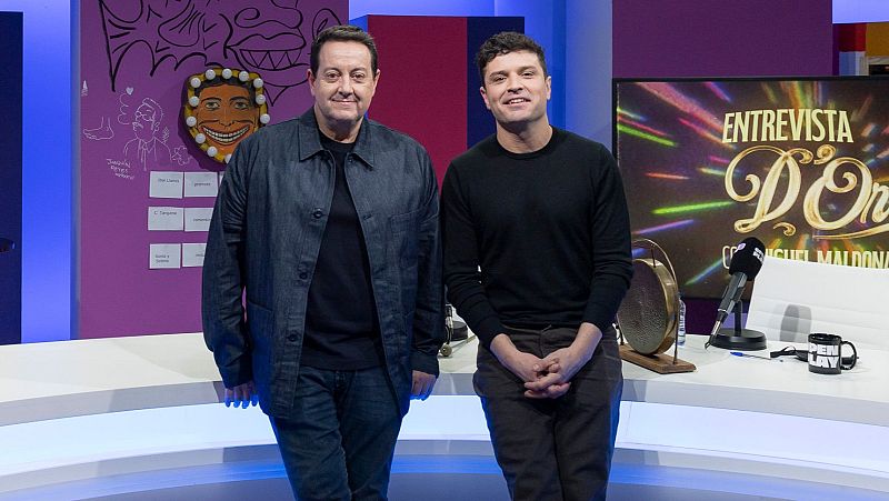 Dos hombres sonríen en un estudio de televisión. Uno viste camisa oscura, chaqueta azul y vaqueros, el otro un jersey negro y pantalones marrones. Un cartel luminoso indica una entrevista.