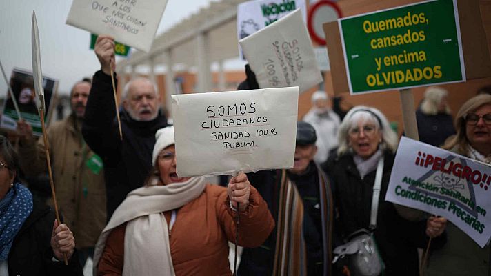 En una manifestación urbana, un grupo de personas exhibe carteles con mensajes manuscritos. Los carteles denuncian la situación de la sanidad pública y las condiciones precarias, reflejando el descontento de los ciudadanos.