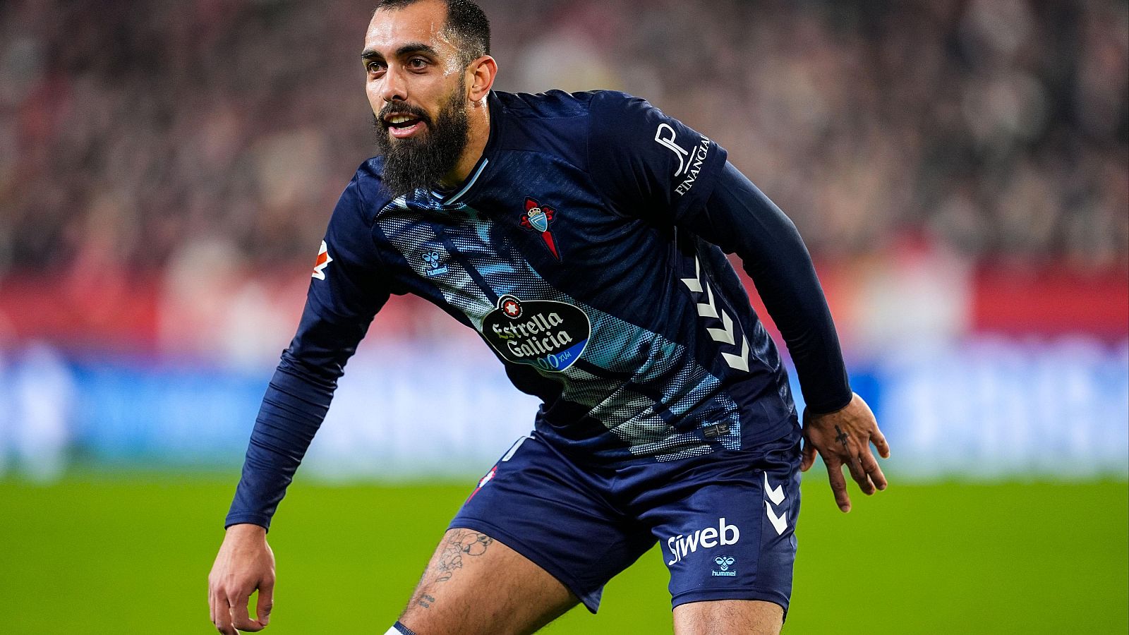 Un futbolista con barba, en un campo de fútbol, luce una camiseta azul oscuro con logos de patrocinadores, incluyendo "Estrella Galicia". Se aprecian tatuajes en sus extremidades.