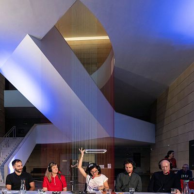 Cinco individuos con auriculares se encuentran sentados en una mesa dentro de un espacio moderno, posiblemente en el IVAM, con vasos de agua y otros objetos. Se observa una instalación artística colgando del techo y otras personas en segundo plano.