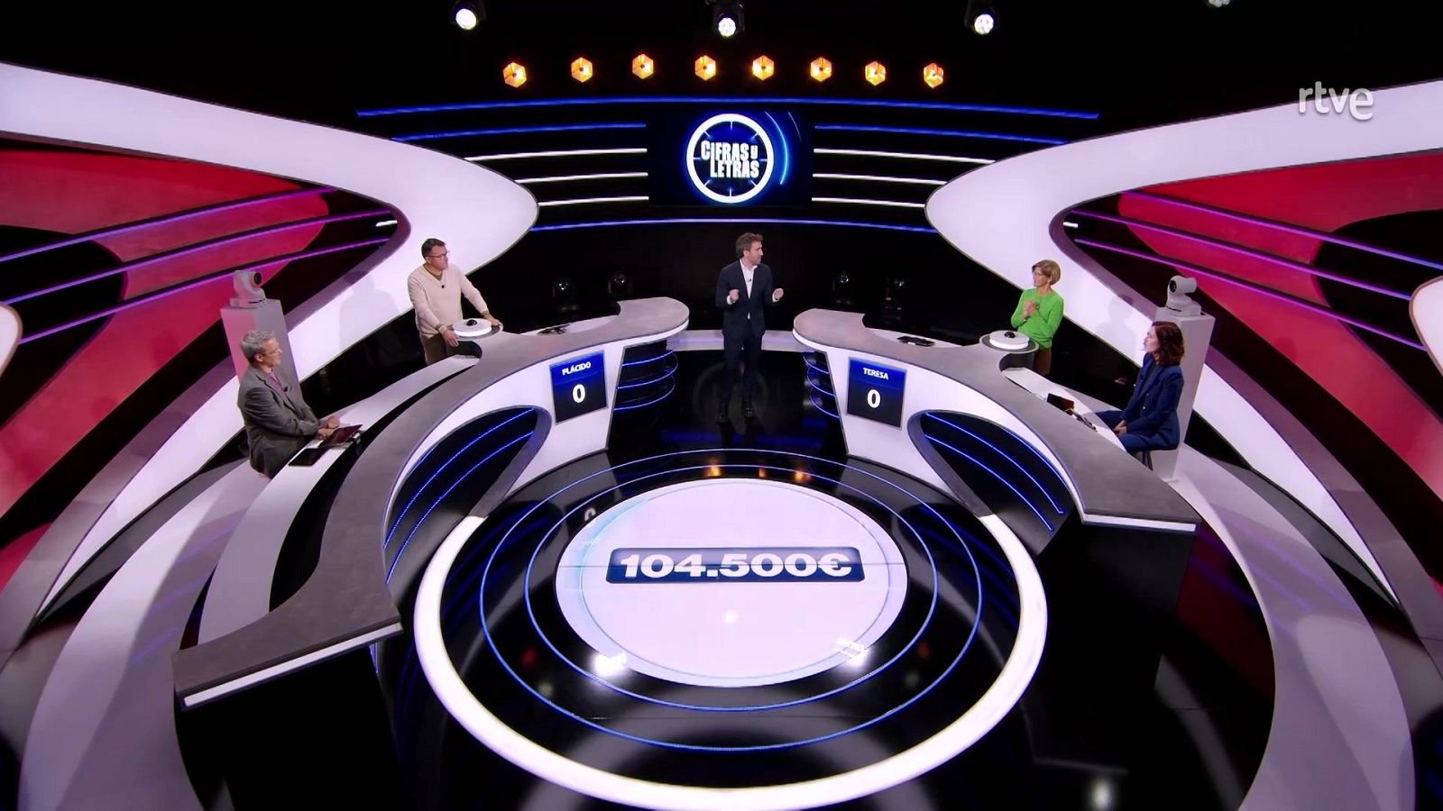 En el plató de un programa de televisión, un presentador se encuentra en el centro, rodeado por concursantes en mesas semicirculares. Se observa un marcador circular con una cantidad y el logo de la cadena en la parte superior.