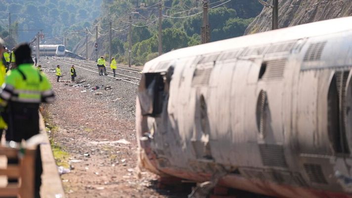 Un tren accidentado con un vagón volcado es el foco principal de la imagen. Personal de seguridad con chalecos reflectantes se encuentra cerca del tren, mientras que otro tren y vegetación se aprecian en el fondo.