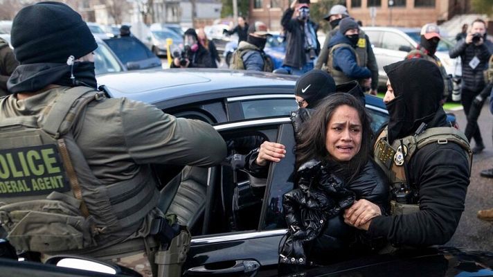 En una calle, una mujer angustiada es asistida por dos personas con chalecos tácticos, mientras un hombre con un chaleco de \"POLICE FEDERAL AGENT\" se encuentra cerca de un coche negro. Otros individuos observan y graban la escena.