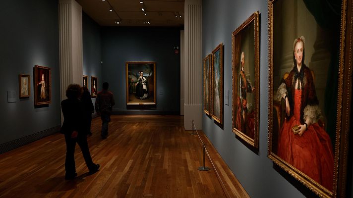 Una sala de museo con suelo de madera y paredes grises exhibe pinturas con marcos dorados. Se observan retratos de figuras, incluyendo una mujer con traje rojo y un chal negro, y dos visitantes contemplando las obras.