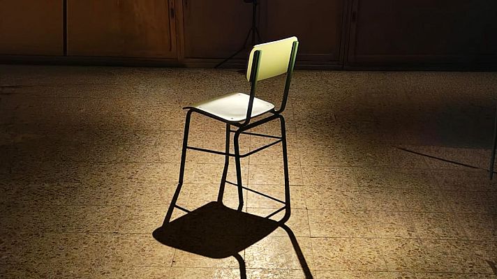 Una silla escolar solitaria, con estructura metálica oscura, asiento blanco y respaldo verde claro, se encuentra en un espacio con suelo de baldosas y una pared oscura, proyectando una sombra alargada.