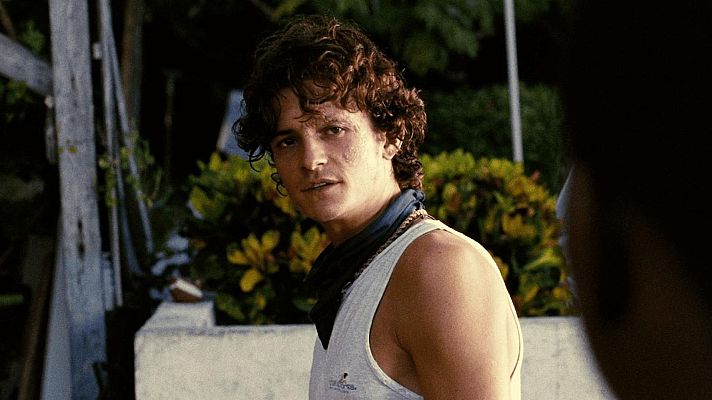 Un hombre joven con cabello rizado y camiseta de tirantes, posa frente a vegetación tropical con flores amarillas, llevando un pañuelo azul oscuro al cuello.