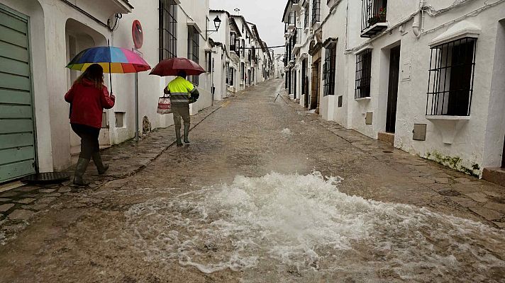 En una calle inundada, dos personas se abren paso bajo la lluvia. Una lleva un paraguas y chaqueta roja, la otra un chaleco reflectante. El agua cubre la calle y emana de una alcantarilla.