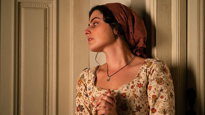 Una mujer joven, con un pañuelo en la cabeza y un vestido floral, se muestra de perfil con una expresión preocupada, con las manos juntas, cerca de una puerta de madera.