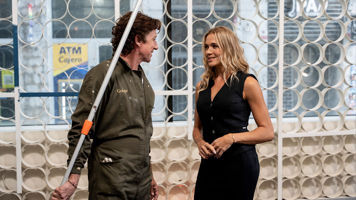 Colate y Paulina Rubio se encuentran en un estudio. Colate viste un mono de trabajo y sostiene un objeto largo, mientras que Paulina Rubio lleva un vestido negro.