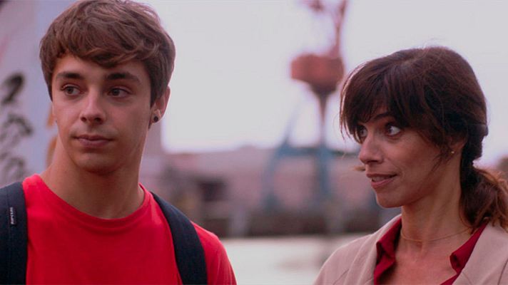 Dos personas, un hombre con camiseta roja y mochila, y una mujer con chaqueta beige, posan mirando hacia la derecha. El fondo muestra una estructura metálica borrosa.