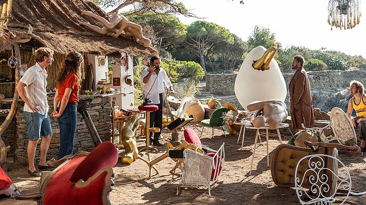 Una escena surrealista al aire libre muestra una colección de muebles, esculturas y un hombre hablando por teléfono. Un huevo gigante con un cisne dorado y una pareja observando la escena completan la composición.