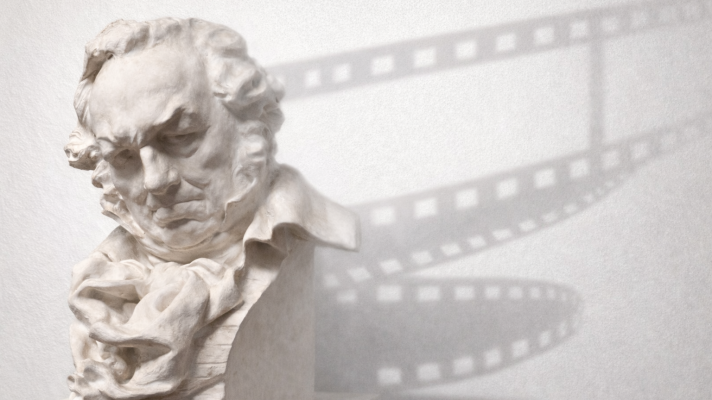 Una escultura de busto blanco representa a un hombre con el ceño fruncido y la mirada baja. Su cabello es rizado y viste una camisa de cuello alto y una chaqueta. Detrás, se proyecta la sombra de una película cinematográfica.