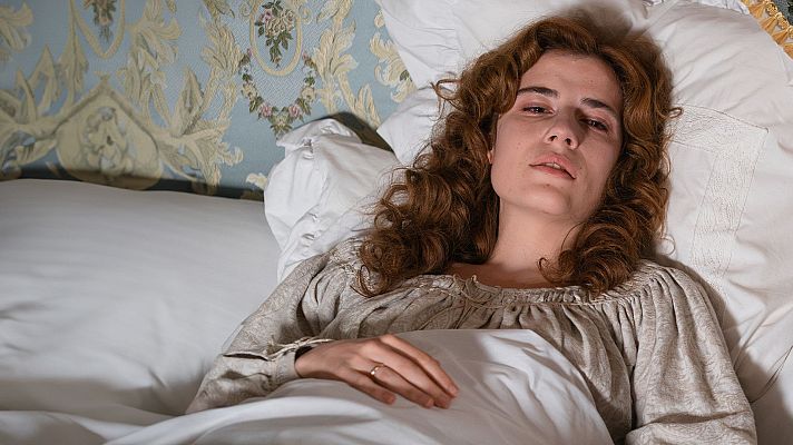 En la imagen, una mujer con cabello rizado y blusa clara descansa en una cama con sábanas y almohadas blancas, y un cabecero con diseño floral.
