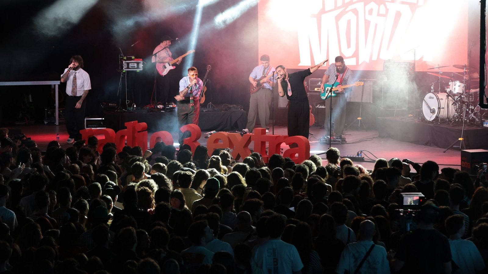 En el escenario, un grupo musical interpreta sus canciones frente a una multitud. Se observan instrumentos como guitarras y batería, y el ambiente es festivo gracias a la iluminación.