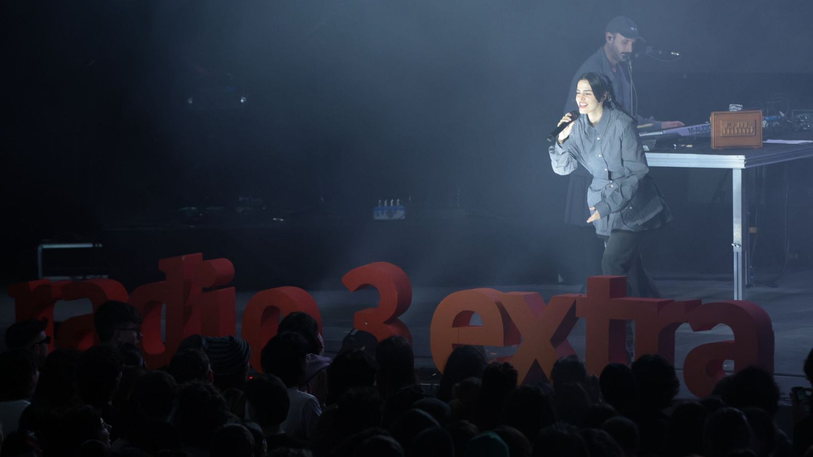 En un concierto con iluminación tenue, una cantante con vestimenta oscura, posiblemente María Arnal, se presenta ante el público, acompañada por un músico que toca el teclado. El escenario está decorado con letras rojas que forman "Radio 3 Extra".