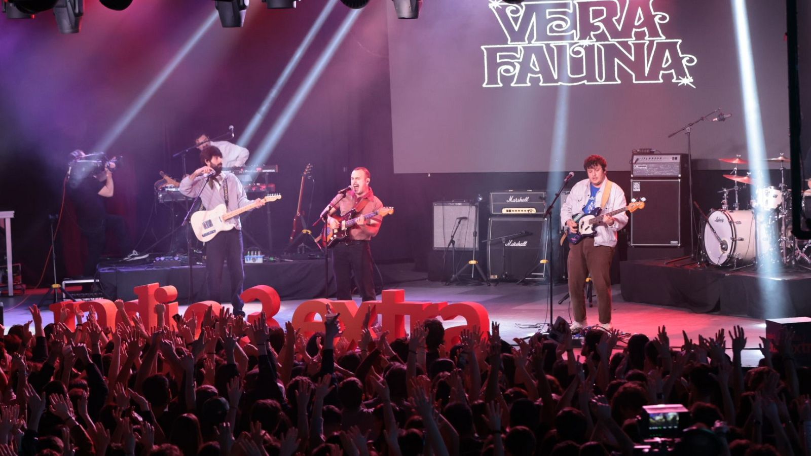 En un escenario iluminado, la banda toca sus instrumentos musicales frente a un público animado. El nombre de la banda, "Vera Fauna", se proyecta en la parte superior del escenario, mientras que "Zona Extra" se lee en la parte inferior.