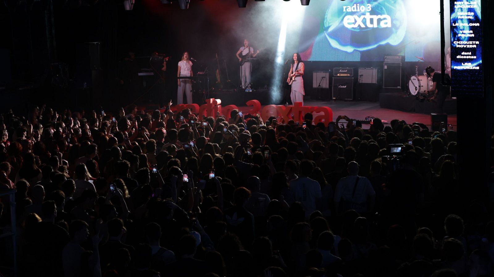 Un grupo musical, con una cantante tocando la guitarra, se presenta en un escenario frente a una multitud. Se observan amplificadores y una pantalla con un logotipo, mientras que el público graba el concierto con sus teléfonos.