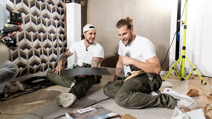 Dos hombres, uno con gorra y tatuajes, trabajan en una instalación, sonriendo. Se encuentran rodeados de materiales de construcción, incluyendo una caja con una marca específica, sobre un suelo de madera.