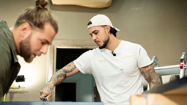 Dos hombres se encuentran trabajando en un espacio interior; uno de ellos, con una camiseta que incluye un nombre y un logo, está inclinado sobre una superficie oscura, mientras que el otro observa la tarea.
