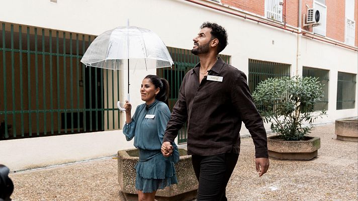 En un patio rodeado de edificios de ladrillo, una pareja camina bajo un paraguas. La mujer, con coleta y vestido azul verdoso, y el hombre, con barba y camisa oscura, se toman de la mano.