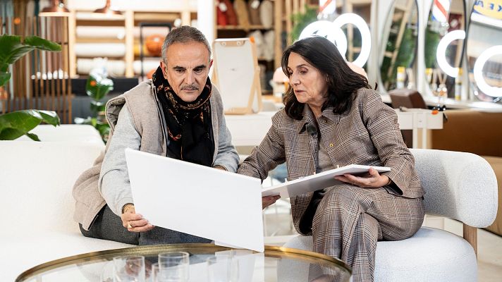 Lorenzo Castillo y Marta Riopérez, en un estudio de diseño, examinan documentos y un portafolio. El espacio, con una mesa de cristal y estanterías con elementos decorativos, sugiere un ambiente dedicado al diseño de interiores.