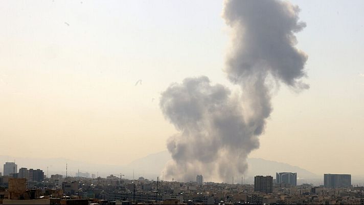 Una ciudad con edificios variados se ve envuelta en una densa columna de humo gris, posiblemente debido a un incidente o explosión. El humo se eleva desde el interior de la ciudad, oscureciendo el cielo.