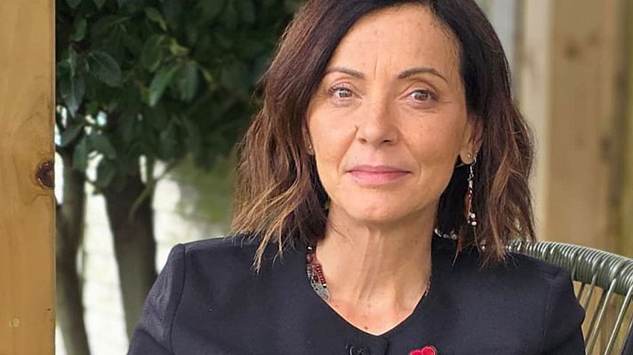En primer plano, una mujer con cabello castaño y ojos marrones posa con una chaqueta negra y accesorios como pendientes y un collar. El fondo, borroso, revela vegetación y elementos de madera.