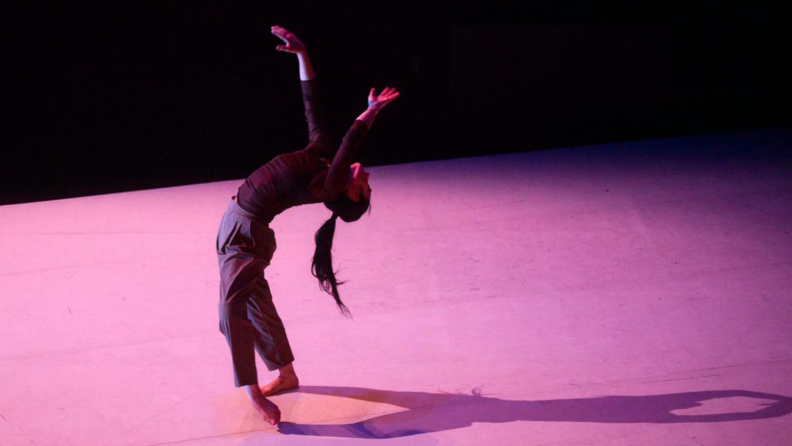 En un escenario con iluminación rosada, una mujer descalza con coleta y vestida con ropa oscura ejecuta un movimiento de danza, arqueando la espalda y levantando los brazos.