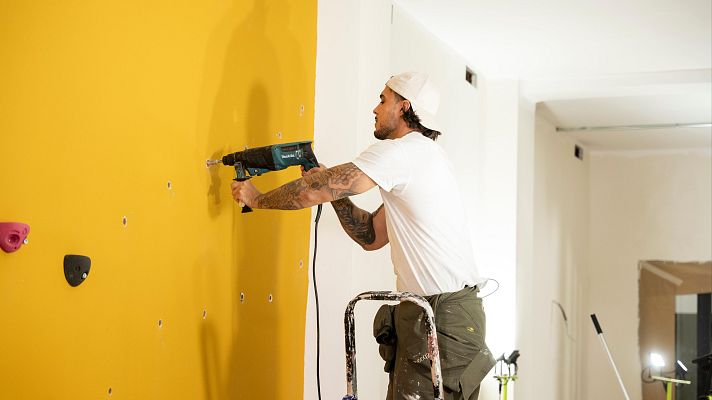 Un hombre con camiseta blanca, pantalones verdes y gorra blanca utiliza un taladro para perforar una pared amarilla con agujeros y agarres de escalada, mientras está de pie sobre una escalera.
