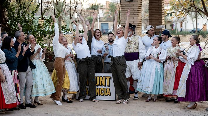 En una celebración al aire libre, un grupo de personas festeja. Un grupo central de cinco personas, vestidas con ropa de trabajo y un trofeo, celebra frente a un cartel. A los lados, personas con trajes tradicionales valencianos.