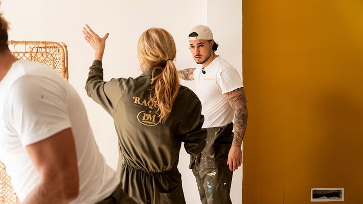 En un set de grabación, un hombre con camiseta blanca y gorra, posiblemente Carlo Constanzia, posa frente a la cámara. Una mujer con un mono de trabajo verde y un hombre con camiseta blanca completan la escena en una habitación en renovación.