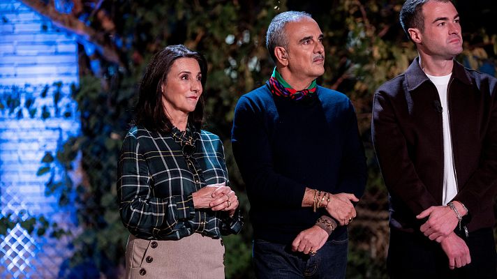 Tres personas posan frente a un fondo con vegetación y luz azul. Una mujer con camisa de cuadros y falda, un hombre con jersey azul y pañuelo, y un hombre joven con chaqueta marrón y camiseta blanca.