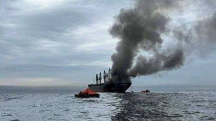 Un barco en llamas genera una columna de humo negro en medio de un mar agitado. Se observan botes salvavidas cerca de la embarcación, uno de ellos de color naranja, bajo un cielo cubierto de nubes grises.