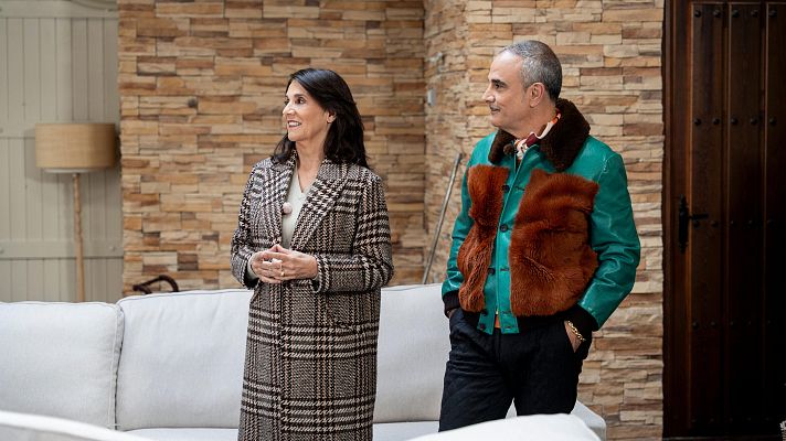 Marta Riopérez y Lorenzo Castillo, vestidos con atuendos elegantes, se encuentran en un espacio interior con una pared de piedra como telón de fondo. Marta lleva un abrigo de cuadros y Lorenzo una chaqueta de cuero, ambos mirando hacia un lado.