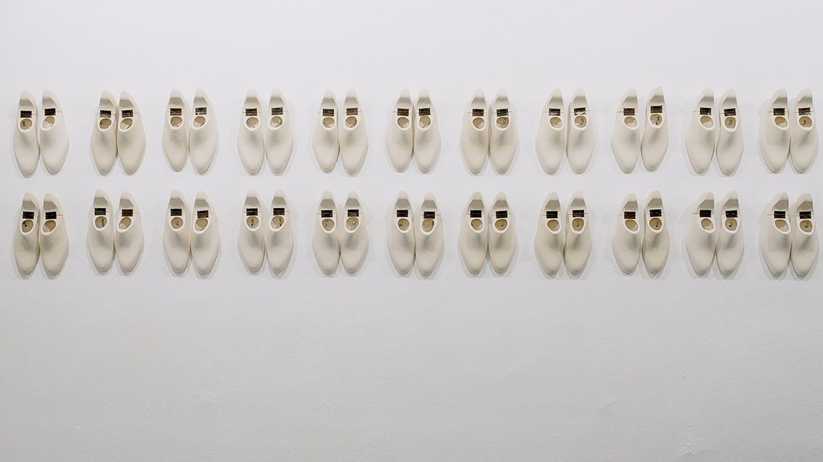 Una fotografía muestra una serie de zapatos blancos idénticos, alineados en filas horizontales sobre un fondo claro, creando una composición simétrica y ordenada.